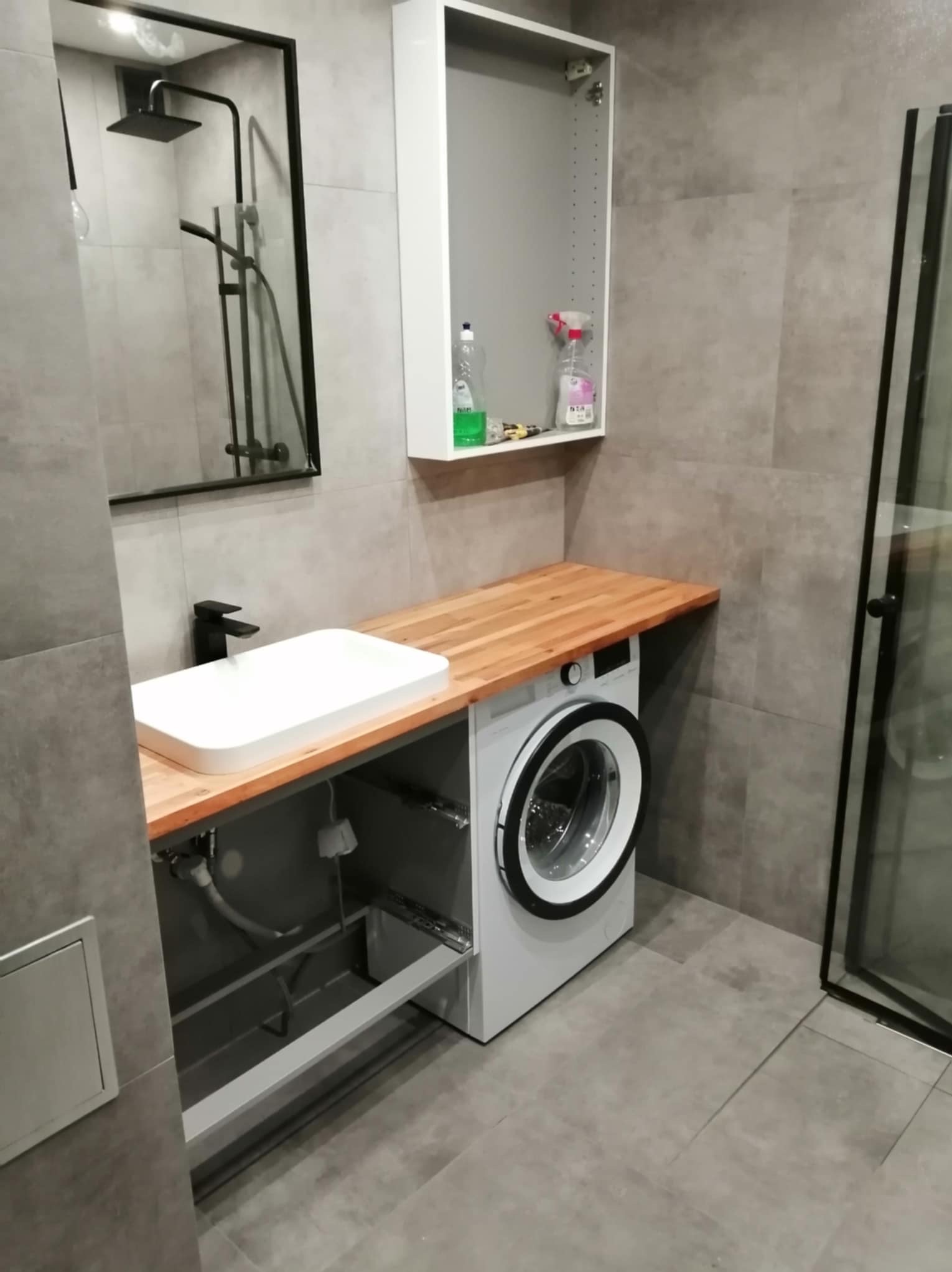 Nowoczesna łazienka w Rybniku z drewnianym blatem, białą umywalką nablatową z czarną armaturą, pralką pod blatem i kabiną prysznicową z czarnymi ramami, widok na wiszącą szafkę z kosmetykami...