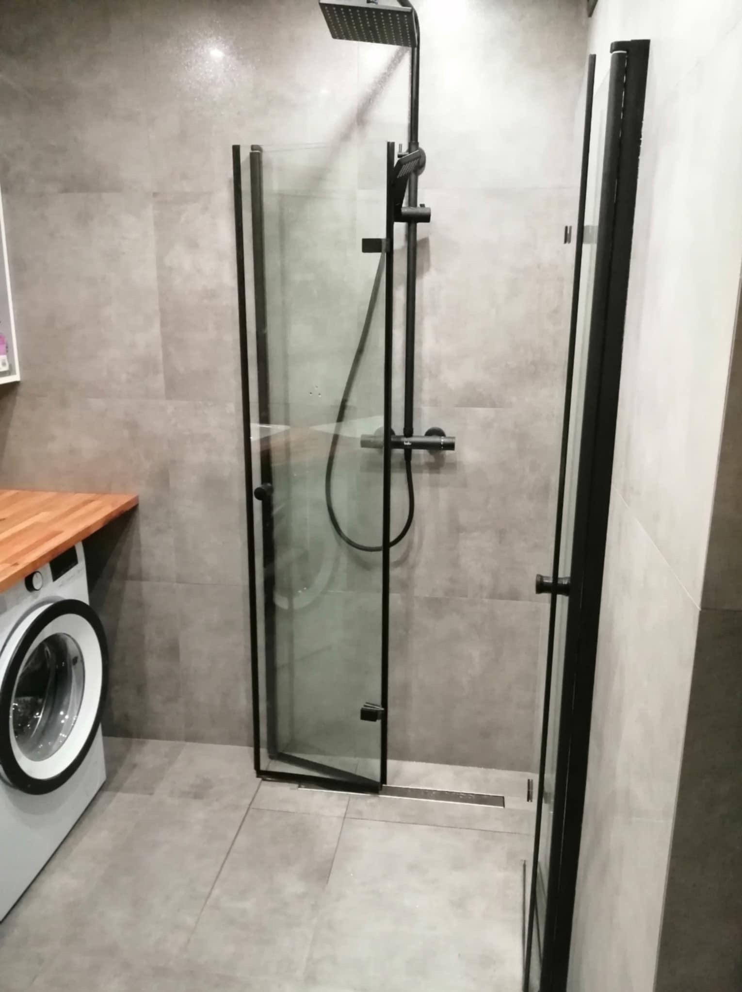 Nowoczesna łazienka z kabiną prysznicową typu walk-in z czarnymi profilami, szara glazura i pralka z drewnianym blatem.