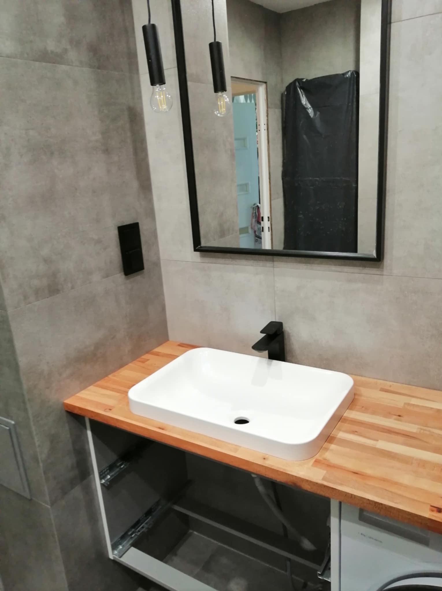 Nowoczesna łazienka w szarościach z drewnianym blatem, białą umywalką nablatową i czarną armaturą, wiszące lampy z żarówkami Edisona, widoczne fragmenty instalacji pod blatem.