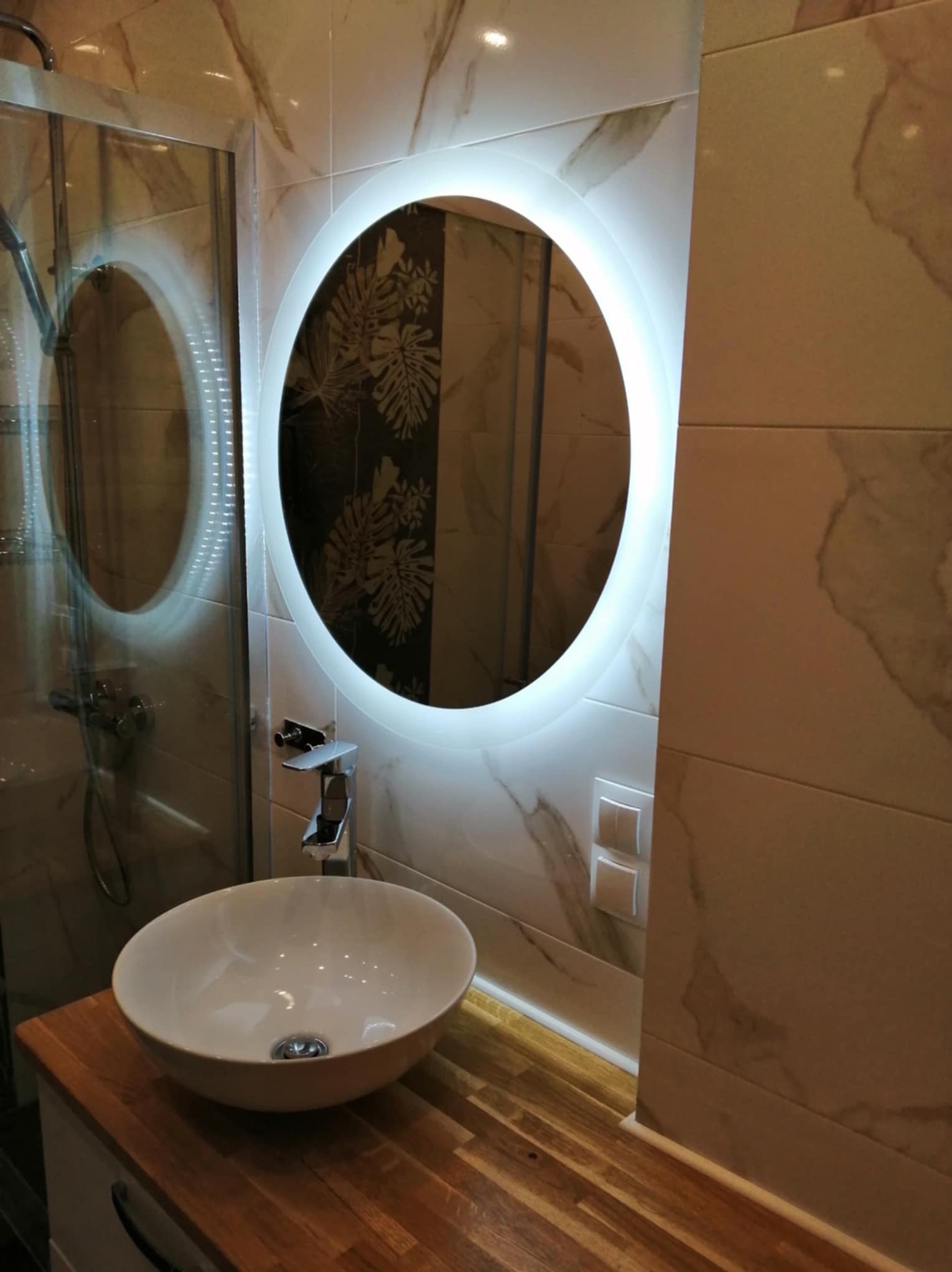 Nowoczesna łazienka z okrągłym lustrem LED, białą umywalką nablatową i drewnianym blatem, kabina prysznicowa w tle.