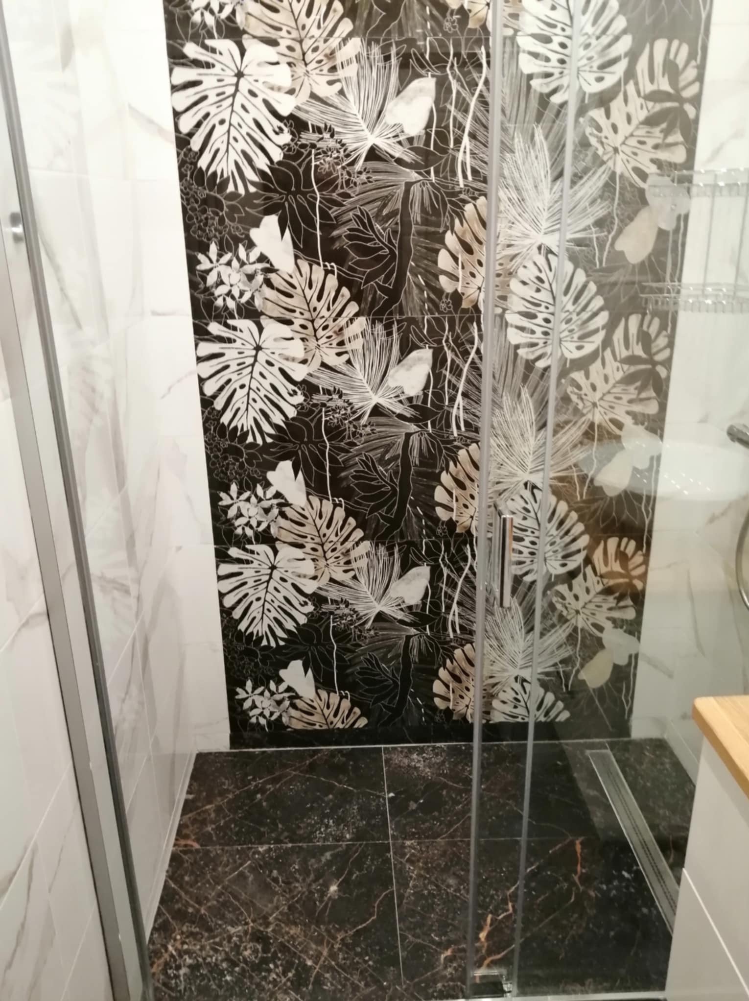 Kabina prysznicowa z czarnymi płytkami w kwiatowe wzory, marmurowa podłoga i białe ściany z efektem marmuru.
