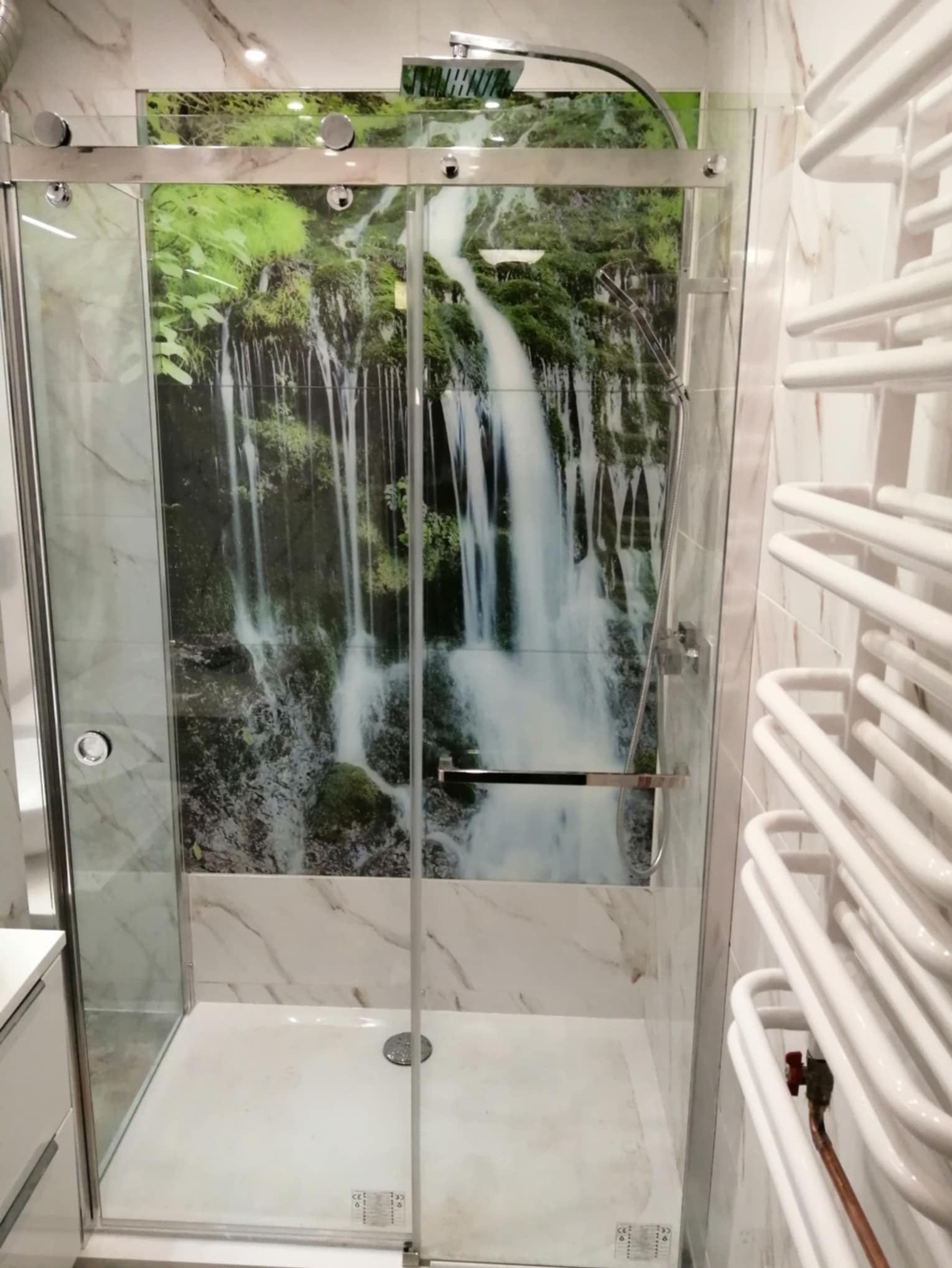 Szklana kabina prysznicowa z motywem wodospadu na tylnej ściance, białe marmurowe płytki na ścianach, biały grzejnik drabinkowy.