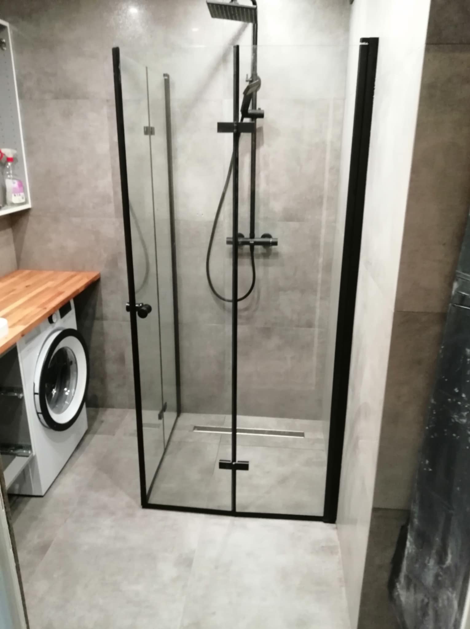 Nowoczesna kabina prysznicowa z czarnymi profilami i deszczownicą, w tle pralka z drewnianym blatem w łazience wykończonej szarymi płytkami.