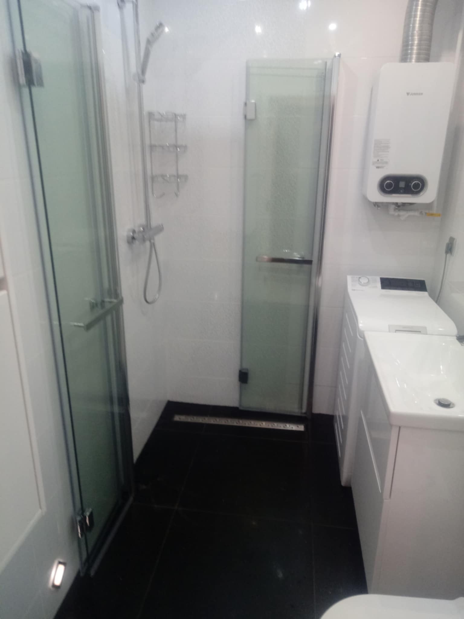 Nowoczesna łazienka z kabiną prysznicową ze szklanymi drzwiami, czarną podłogą, białą umywalką z szafką, pralką i piecem gazowym na ścianie.