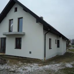 Nowoczesny dom jednorodzinny z jasną, gładką elewacją, ciemnym dachem i brązowymi oknami. Widoczne elementy wykończeniowe, takie jak rynny i balkon. Teren wokół domu częściowo pokryty śniegiem.