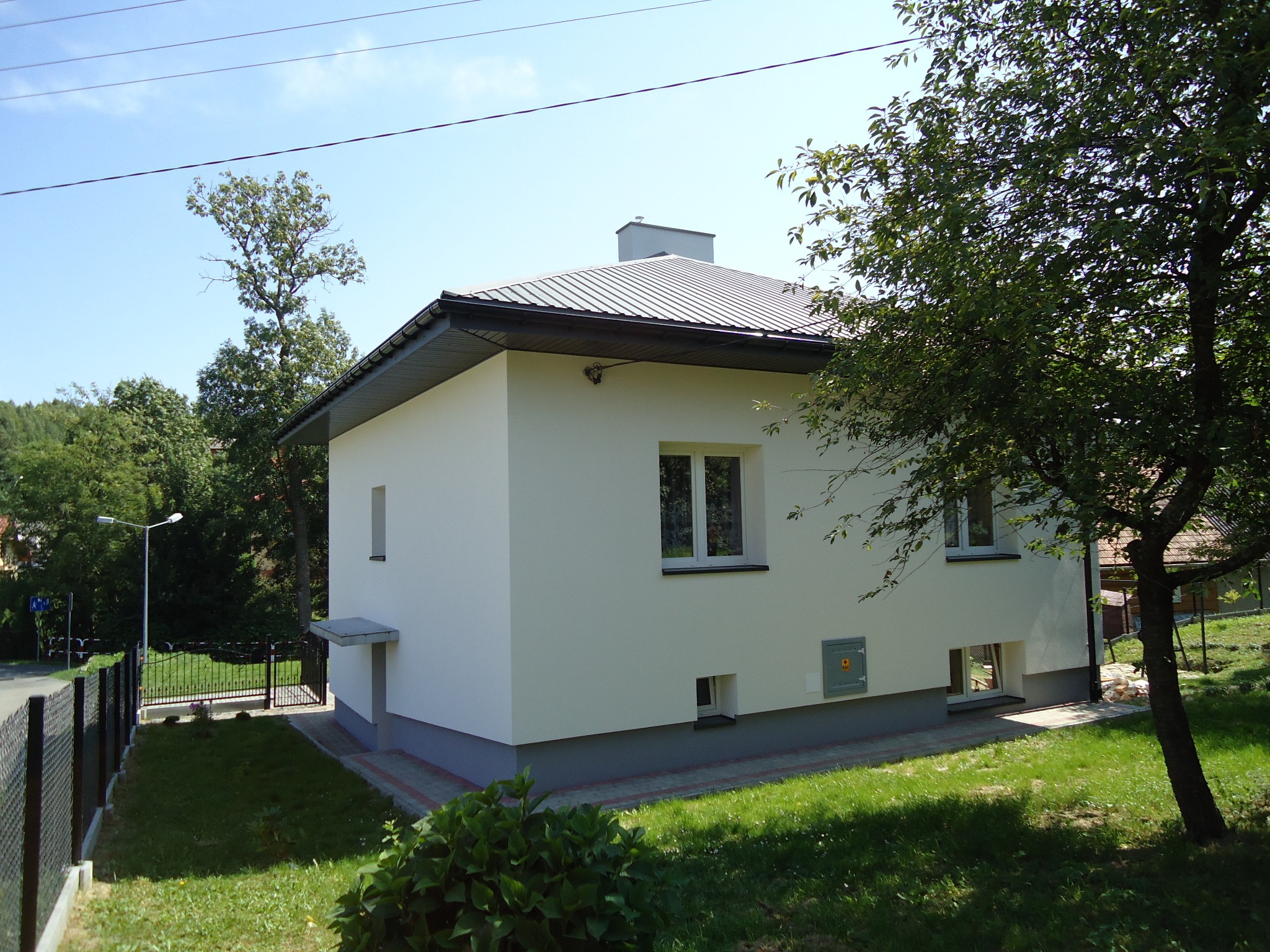 Biały, otynkowany dom jednorodzinny z szarym dachem i elementami wykończeniowymi, widoczny z perspektywy ogrodu w słoneczny dzień. Na elewacji widoczne okna z białymi ramami i skrzynka gazowa.