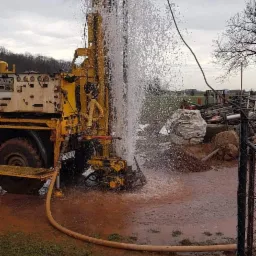 Żółta maszyna wiertnicza w trakcie pracy, z silnym strumieniem wody tryskającym spod ziemi, teren wokół maszyny błotnisty, w tle widoczne drzewa i pochmurne niebo.