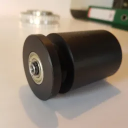 Czarny, matowy wałek z łożyskiem kulkowym na krawędzi, obok widoczny pasek zębaty i segregator biurowy w tle.