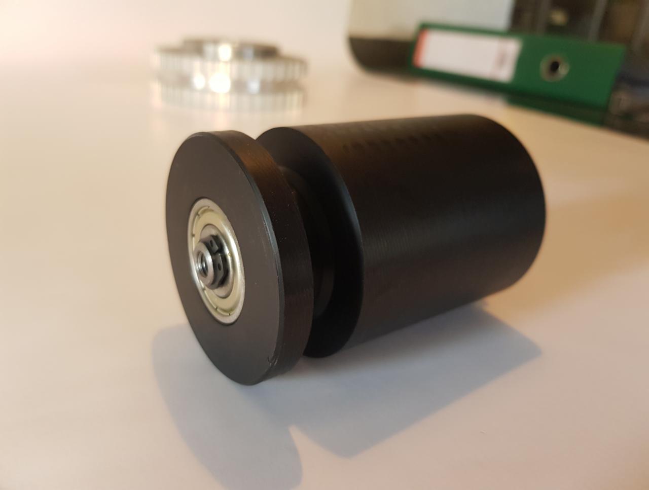 Czarny, matowy wałek z łożyskiem kulkowym na krawędzi, obok widoczny pasek zębaty i segregator biurowy w tle.