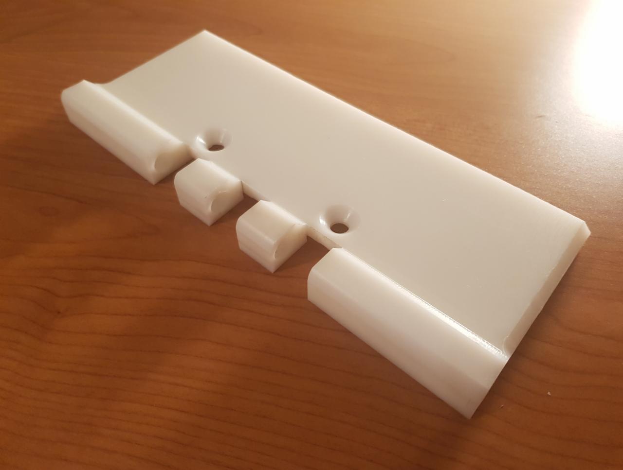 Biały element z tworzywa sztucznego z dwoma otworami i wypustkami, leżący na drewnianym blacie, widoczne ślady obróbki.