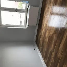 Świeżo położona, błyszcząca podłoga z paneli w odcieniach brązu, widok z góry, szare ściany, białe listwy przypodłogowe i okno z widokiem na zewnątrz.