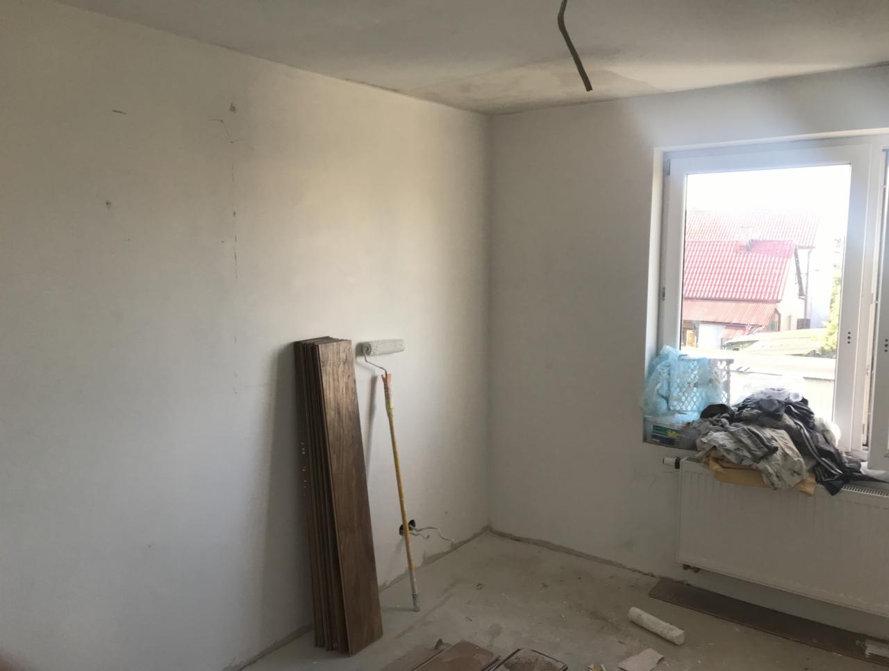 Pokój w trakcie prac przygotowawczych: świeżo pomalowane ściany na biało, paczka paneli podłogowych oparta o ścianę obok wałka do malowania, okno z widokiem na zabudowę jednorodzinną.