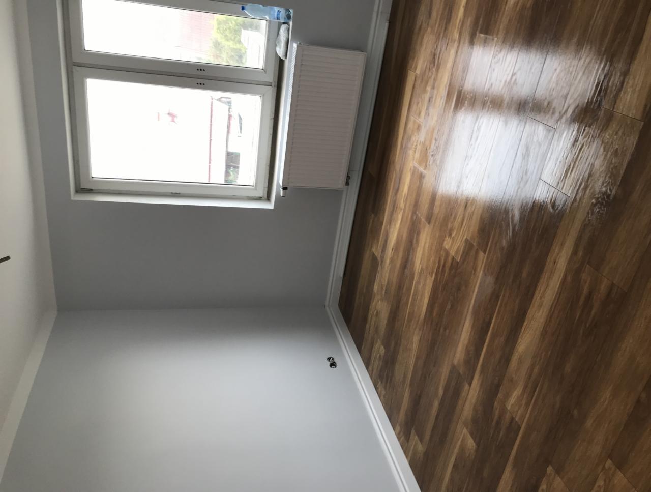 Świeżo położona, błyszcząca podłoga z paneli w odcieniach brązu, widok z góry, szare ściany, białe listwy przypodłogowe i okno z widokiem na zewnątrz.