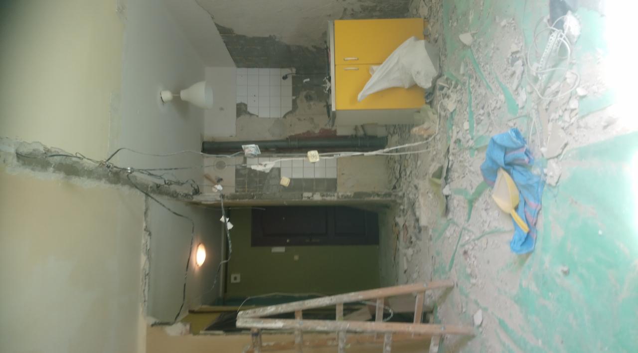 Wnętrze w trakcie demontażu: zerwane płytki, odsłonięte przewody, żółta szafka, gruz na podłodze, widoczna drabina.