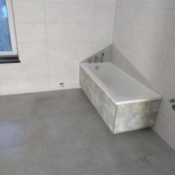 Usługi ogólnobudowlane Damian Ciemny - Wykończona łazienka z szarymi płytkami podłogowymi, wanną obudowaną płytkami imitującymi kamień i białymi płytkami ściennymi, widoczne niedokończone mocowanie na ścianie.