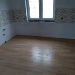 Ułożona podłoga z paneli imitujących drewno w pomieszczeniu z częściowo wykończoną mozaiką na ścianie i oknem.