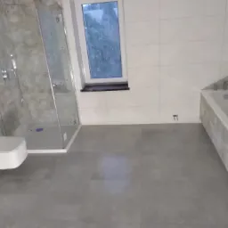 Wykończona łazienka z szarymi płytkami podłogowymi i ściennymi, białą wanną oraz kabiną prysznicową z przezroczystego szkła, widok na okno z czarnym parapetem.