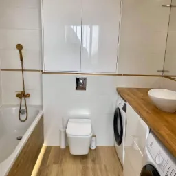 Nowoczesna łazienka po renowacji: białe płytki z złotymi akcentami, drewnopodobna podłoga i obudowa wanny, umywalka nablatowa na drewnianym blacie, zabudowa nad toaletą i dwie pralki.