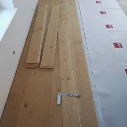 Przygotowanie do ułożenia drewnianej podłogi: deski ułożone obok siebie, podkład zabezpieczający, kątownik pomiarowy. Widoczne niedoskonałości w strukturze drewna.