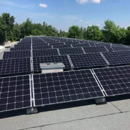 Rząd paneli słonecznych zamontowanych na płaskim dachu, widoczne betonowe podstawy, w tle zieleń drzew i błękitne niebo z chmurami.