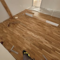 Dariusz Kamiński Kampol - Fragment pokoju z częściowo ułożonym parkietem dębowym, widoczne narzędzia pomiarowe, elementy podkładu podłogowego i fragment nogawki spodni roboczych w dolnym rogu kadru.