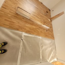 Dariusz Kamiński Kampol - Montaż parkietu w pomieszczeniu: widoczne fragmenty ułożonej podłogi, paczki parkietu oraz zabezpieczona folią ściana. W rogu kadru widoczne buty montera.