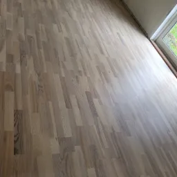 Nowo ułożony parkiet z wyraźnym usłojeniem drewna, widok na fragment ściany z oknem, pomieszczenie w trakcie wykańczania.