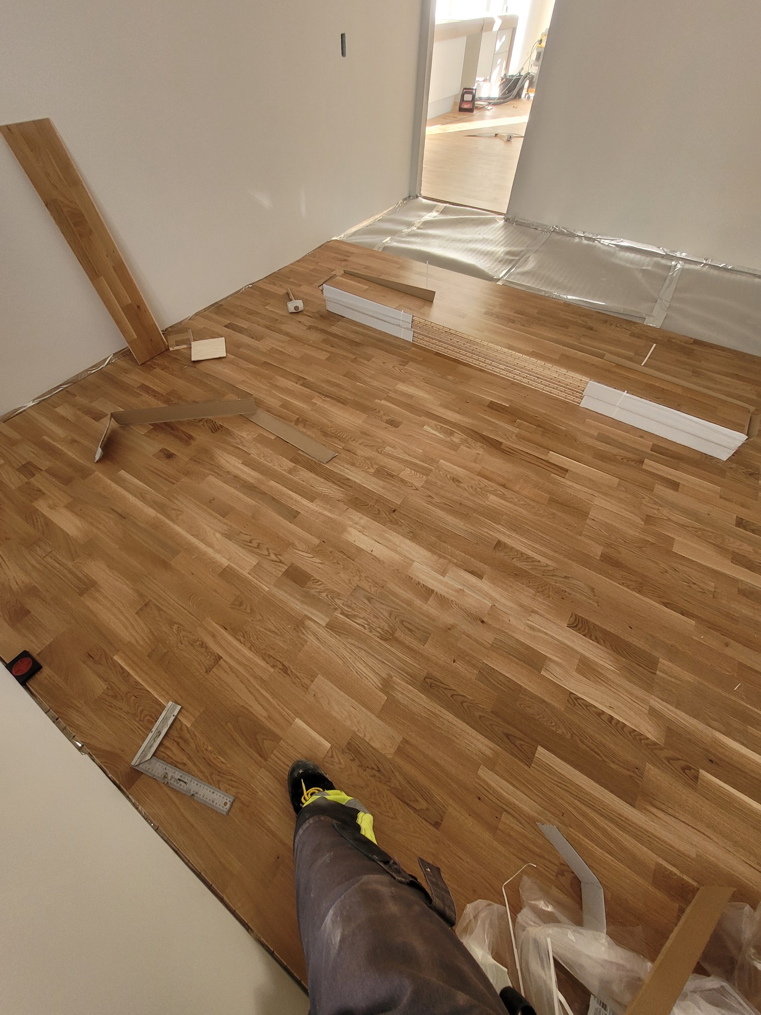 Fragment pokoju z częściowo ułożonym parkietem dębowym, widoczne narzędzia pomiarowe, elementy podkładu podłogowego i fragment nogawki spodni roboczych w dolnym rogu kadru.
