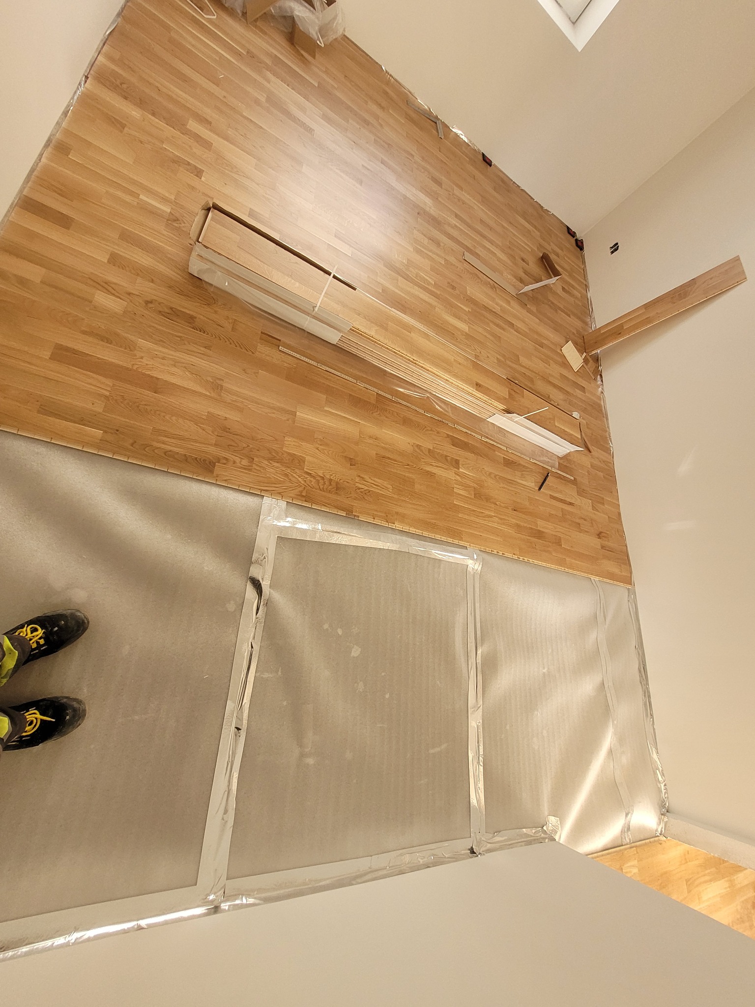 Montaż parkietu w pomieszczeniu: widoczne fragmenty ułożonej podłogi, paczki parkietu oraz zabezpieczona folią ściana. W rogu kadru widoczne buty montera.