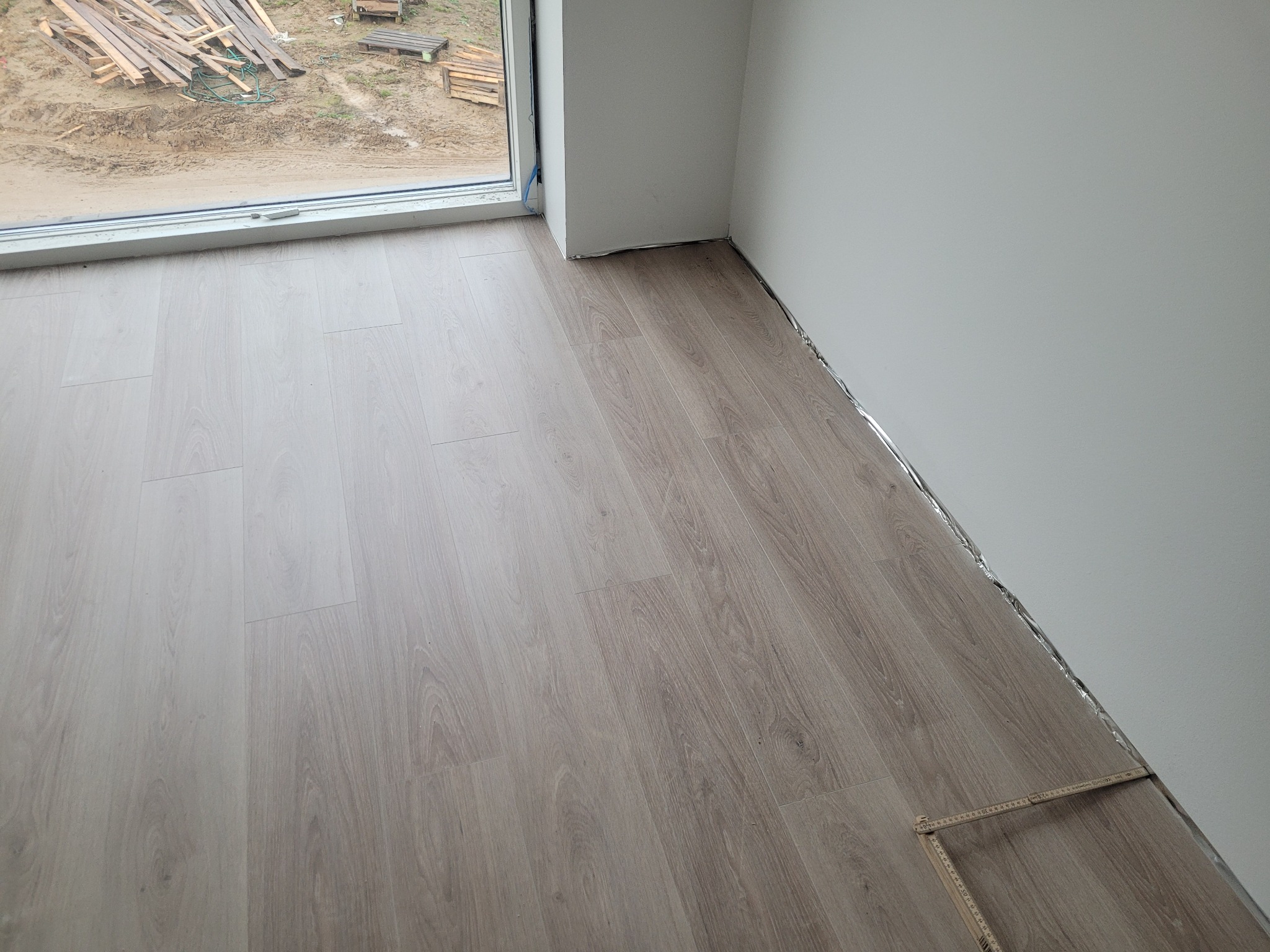 Jasne panele podłogowe ułożone w rogu pokoju, widoczna listwa przypodłogowa, za oknem stos drewna i błoto.