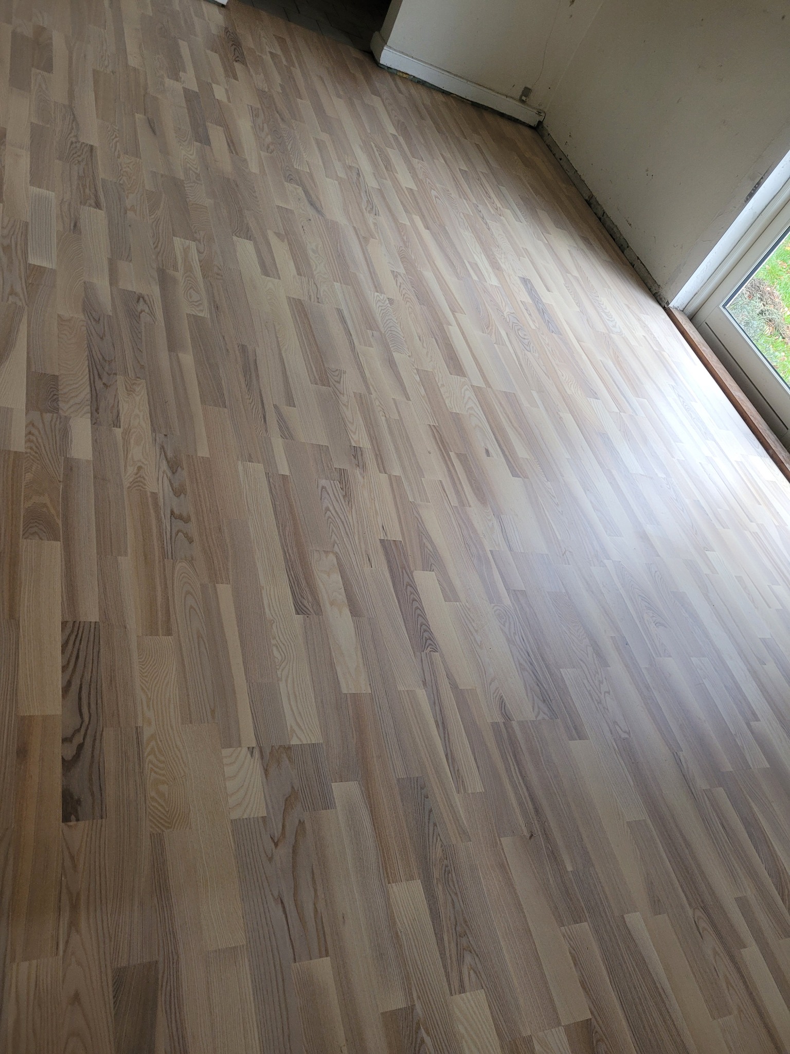 Nowo ułożony parkiet z wyraźnym usłojeniem drewna, widok na fragment ściany z oknem, pomieszczenie w trakcie wykańczania.