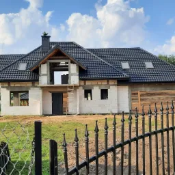 Niedokończony, parterowy dom z czarną dachówką i widoczną konstrukcją dachu, sfotografowany zza metalowego ogrodzenia z ozdobnymi grotami, na tle błękitnego nieba z chmurami.