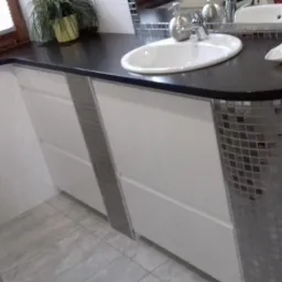 Łazienka z umywalką nablatową na czarnym blacie z białymi szafkami zdobionymi srebrnymi mozaikami, widok z boku na zaokrąglony fragment blatu.