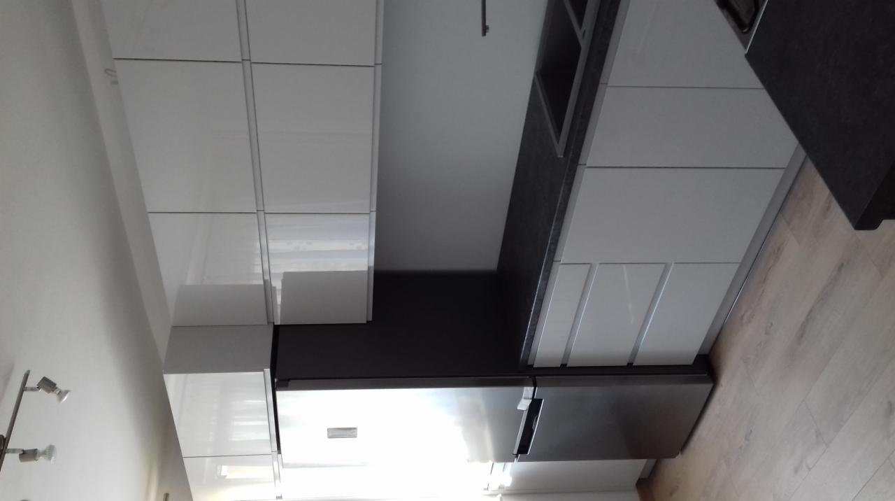 Nowoczesna kuchnia z białymi, lakierowanymi frontami szafek, ciemnym blatem i zabudowaną lodówką ze stali nierdzewnej, oświetlona punktowymi lampami.