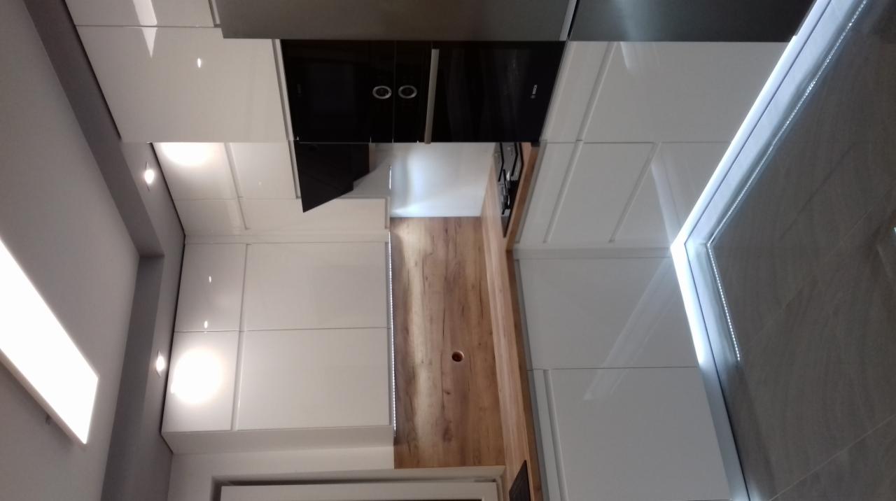 Nowoczesna kuchnia z białymi, lakierowanymi frontami i drewnianym panelem ściennym z oświetleniem LED, widoczny czarny piekarnik w zabudowie.