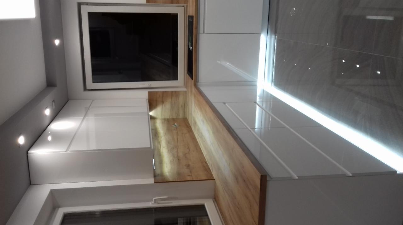 Nowoczesna kuchnia z białymi szafkami i drewnianym blatem, widok z dołu, podkreślony przez oświetlenie punktowe i błyszczące płytki podłogowe.