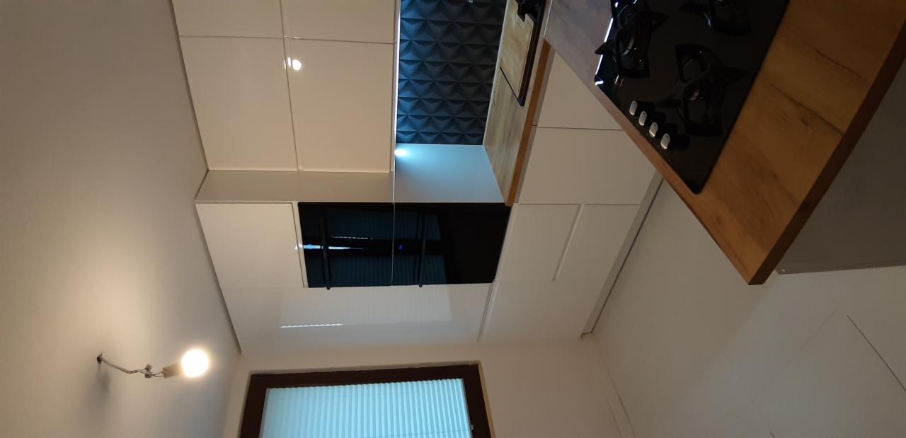 Nowoczesna kuchnia z białymi, błyszczącymi frontami szafek, drewnianym blatem i czarną płytą grzewczą, uzupełniona trójwymiarową ścianą w tle.