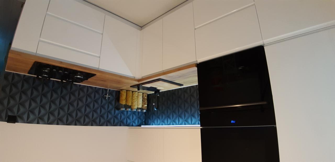 Nowoczesna kuchnia z białymi szafkami, drewnianym blatem, czarną płytą grzewczą i ścianą z trójwymiarowych paneli; widoczne pojemniki na produkty sypkie.