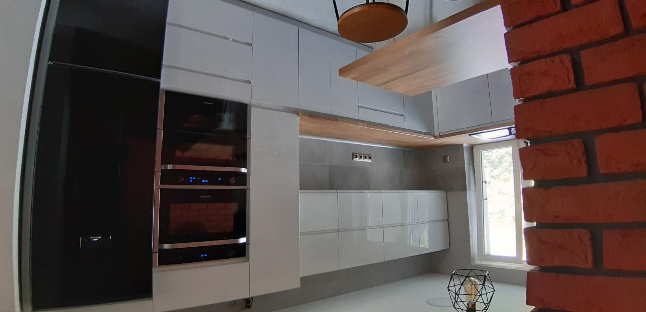 Nowoczesna kuchnia z białymi, błyszczącymi szafkami, zabudowanym piekarnikiem i kuchenką mikrofalową, drewnianym blatem, szarą ścianą i dekoracyjną lampą w kształcie geometrycznej kuli.