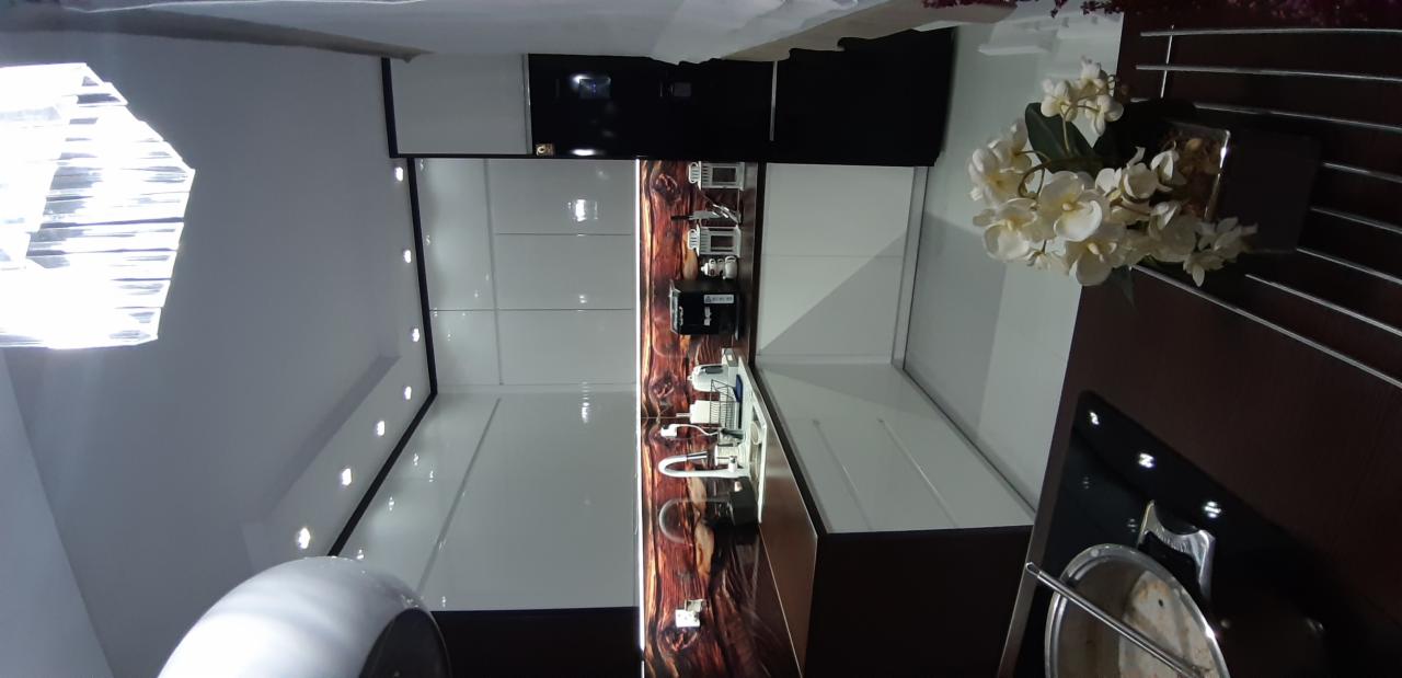 Nowoczesna, biała kuchnia z połyskiem, z akcentem dekoracyjnej ściany imitującej drewno, oświetleniem punktowym i kryształowym żyrandolem, uzupełniona kwiatami w doniczce.