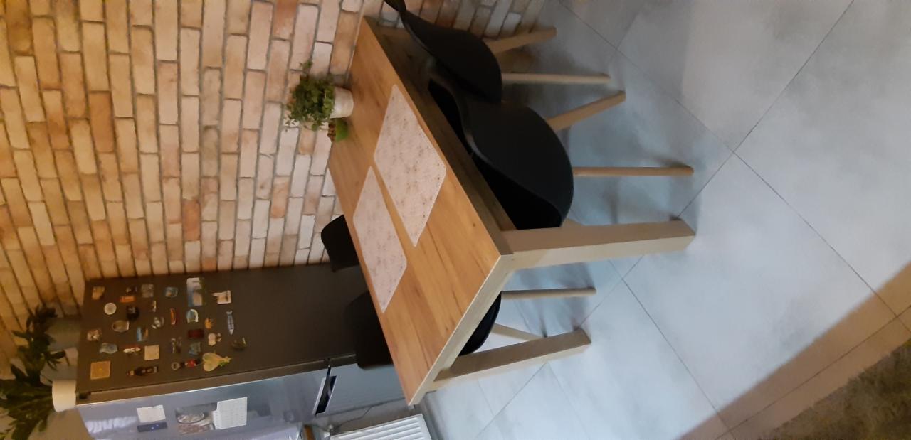 Drewniany stół z jasnymi nogami i czarnymi krzesłami na tle ceglanej ściany i lodówki z magnesami.