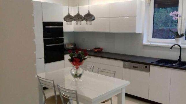 Biała kuchnia z połyskiem, czarny blat, chromowane lampy wiszące i stół z kwiatami w wazonie, widok na okno z rośliną.