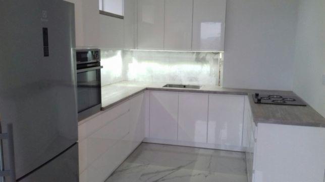 Nowoczesna, biała kuchnia z połyskiem, szary blat, srebrna lodówka i marmurowa podłoga.