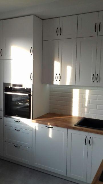 Jasna kuchnia z białymi szafkami, drewnianym blatem i czarnymi uchwytami, oświetlona popołudniowym słońcem padającym na fronty szafek i białe kafelki.