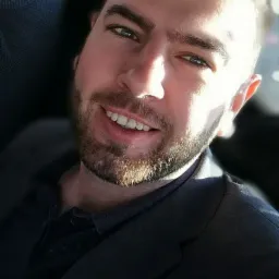 Portret uśmiechniętego mężczyzny z kr&oacute;tką brodą, ubranego w ciemną marynarkę, z ostrym światłem padającym na twarz i widoczną częścią szyldu w tle.