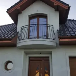 Elewacja domu jednorodzinnego z balkonem z czarną, ozdobną balustradą i drewnianymi oknami oraz drzwiami wejściowymi z owalnym przeszkleniem.