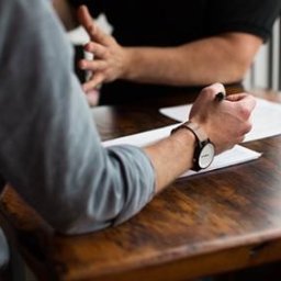 Kancelaria Prawna Solace - Spotkanie dwóch osób przy drewnianym stole. Jedna osoba trzyma długopis nad dokumentami, druga gestykuluje. Widoczne zegarek na ręce i fragment okna w tle.