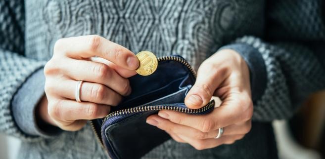 Osoba wkłada monetę do otwartego, granatowego portfela. Widoczne dłonie z pierścionkami i fragment wzorzystego swetra. Kontekst finansowy, oszczędności.