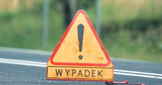 Żółty znak ostrzegawczy z wykrzyknikiem i napisem 'WYPADEK' na tle asfaltowej drogi i zieleni. Znak lekko zniszczony, ustawiony na poboczu.