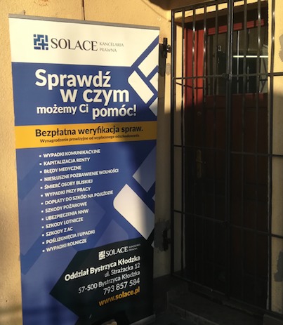 Baner reklamowy Kancelarii Prawnej Solace z ofertą bezpłatnej weryfikacji spraw, umieszczony obok wejścia do budynku. Widoczne dane kontaktowe i lista spraw.